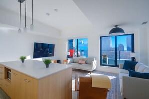 Ein modernes Wohnzimmer mit großem Fernseher, einer Couch, einem Couchtisch und Blick auf die Stadt durch große Fenster.