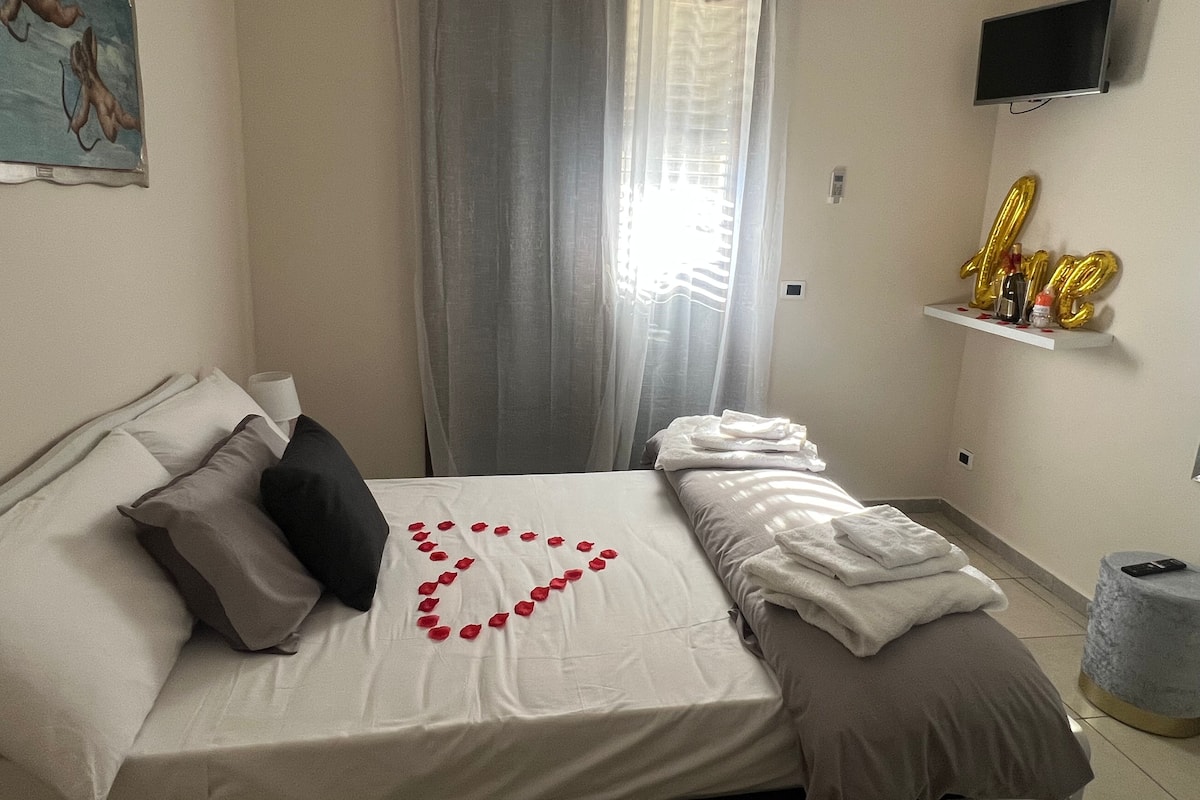 A neatly made bed with a heart-shaped arrangement of rose petals, a television mounted on the wall, and a shelf with golden balloons spelling "LOVE".