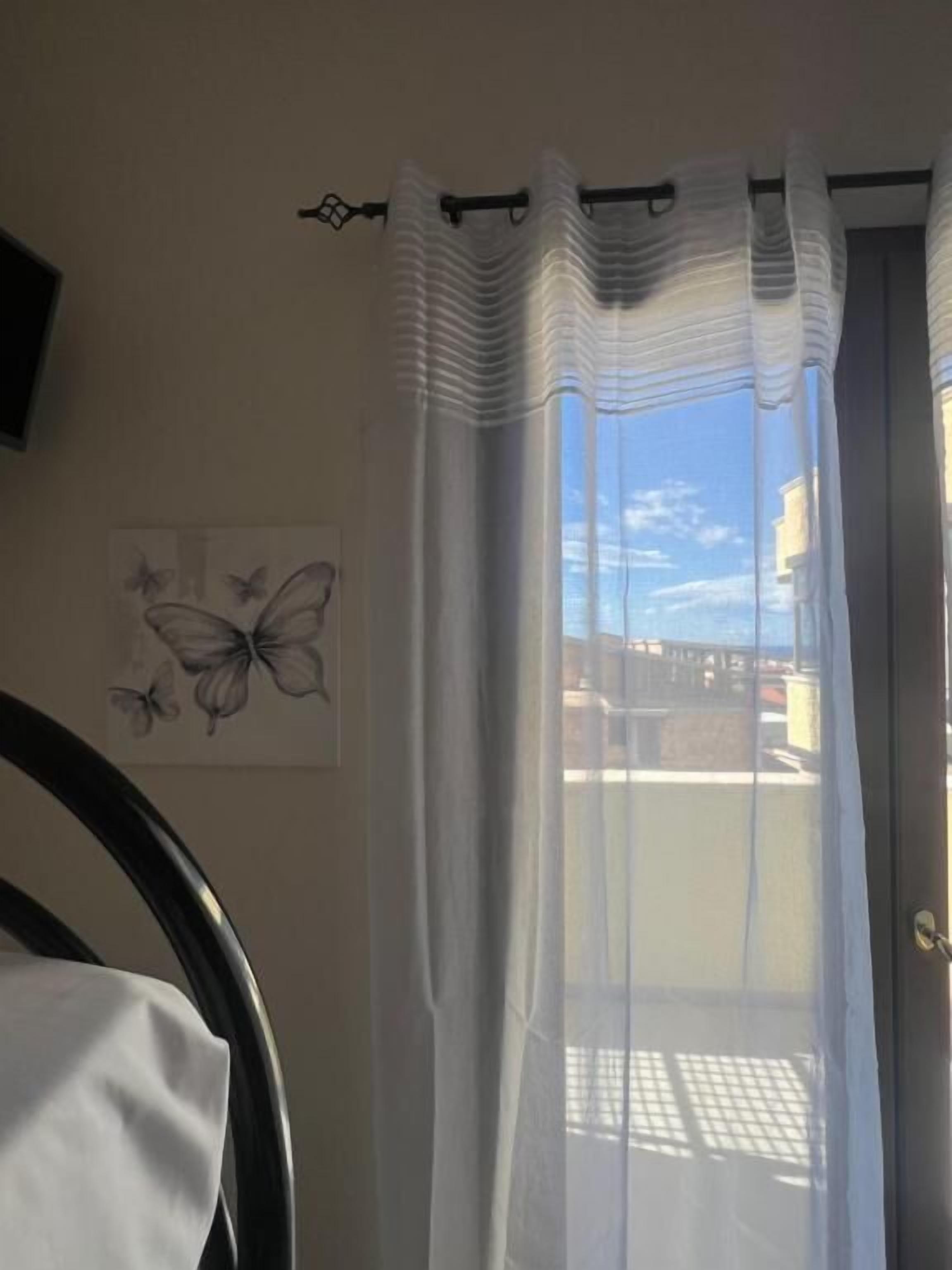 A room with a view of a building through a window with sheer curtains. There is a door with a metal handle and a wall-mounted light fixture.