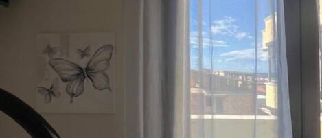A room with a view of a building through a window with sheer curtains. There is a door with a metal handle and a wall-mounted light fixture.