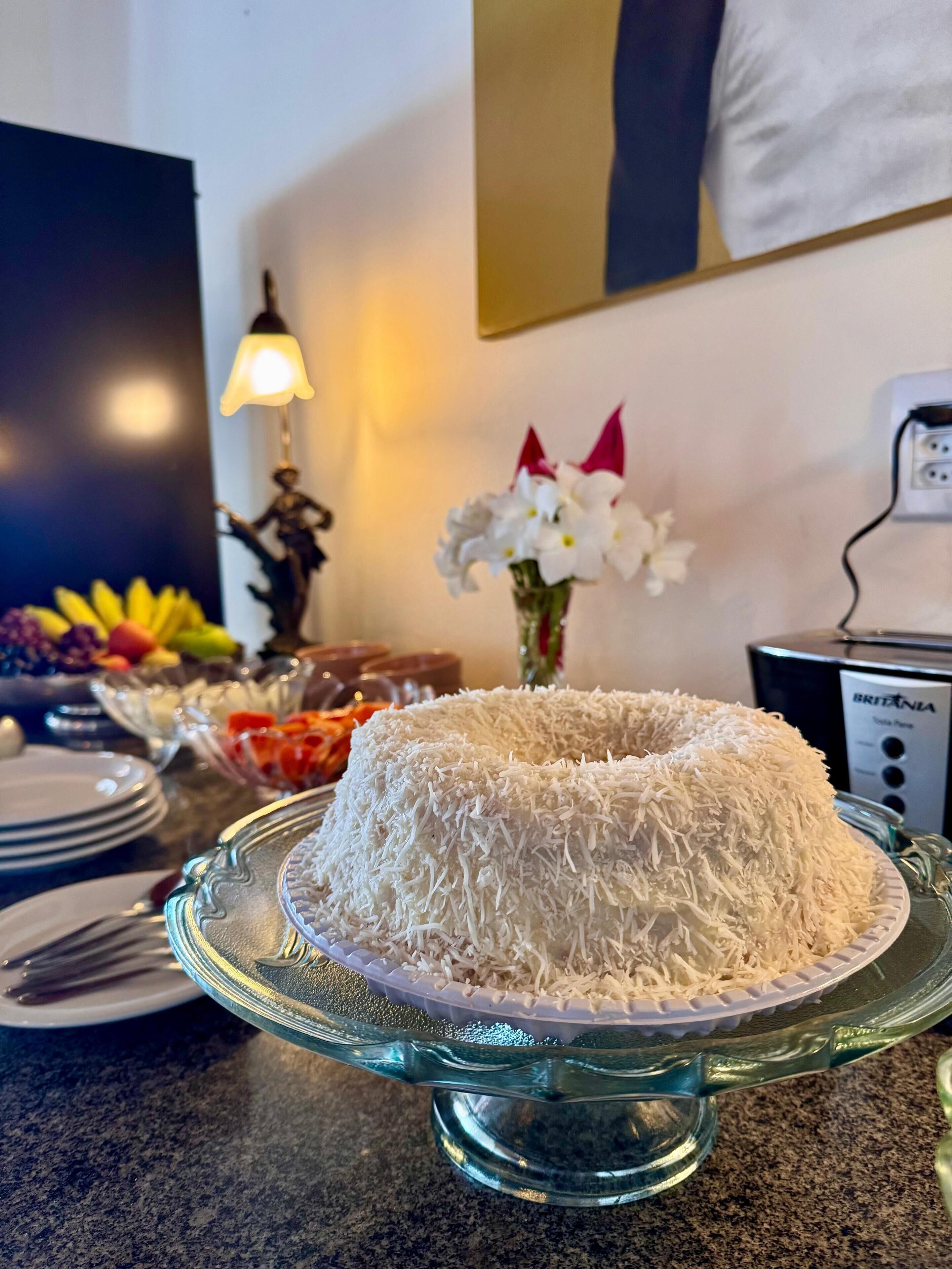 A glass cake stand with a coconut-covered cake on a granite countertop.