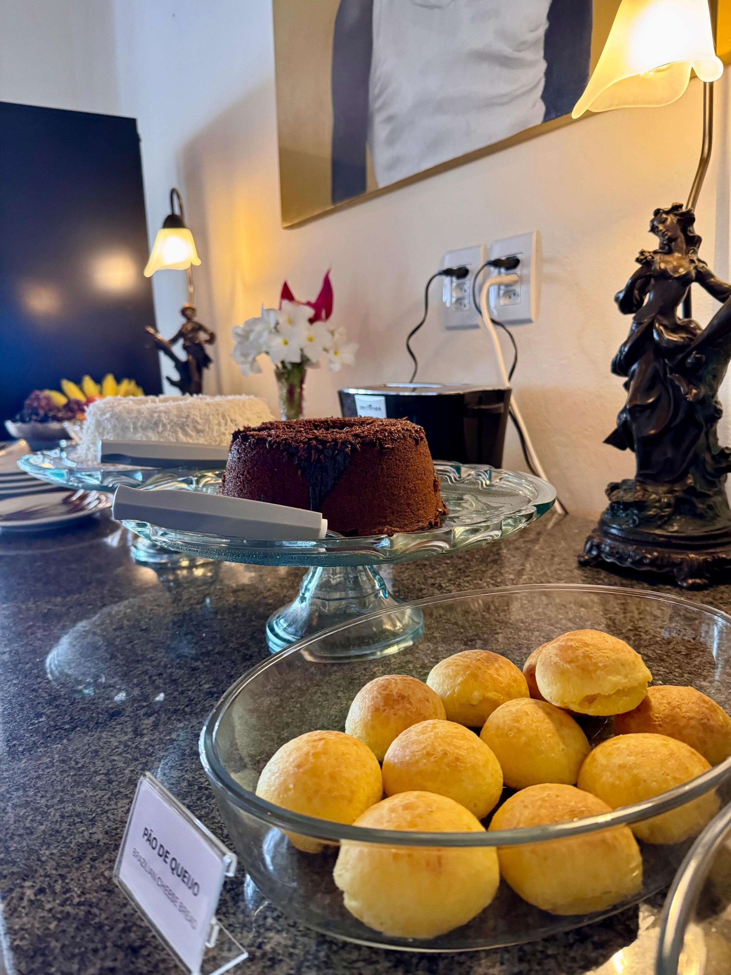 A dessert display with a chocolate cake and pão de queijo on a glass stand.