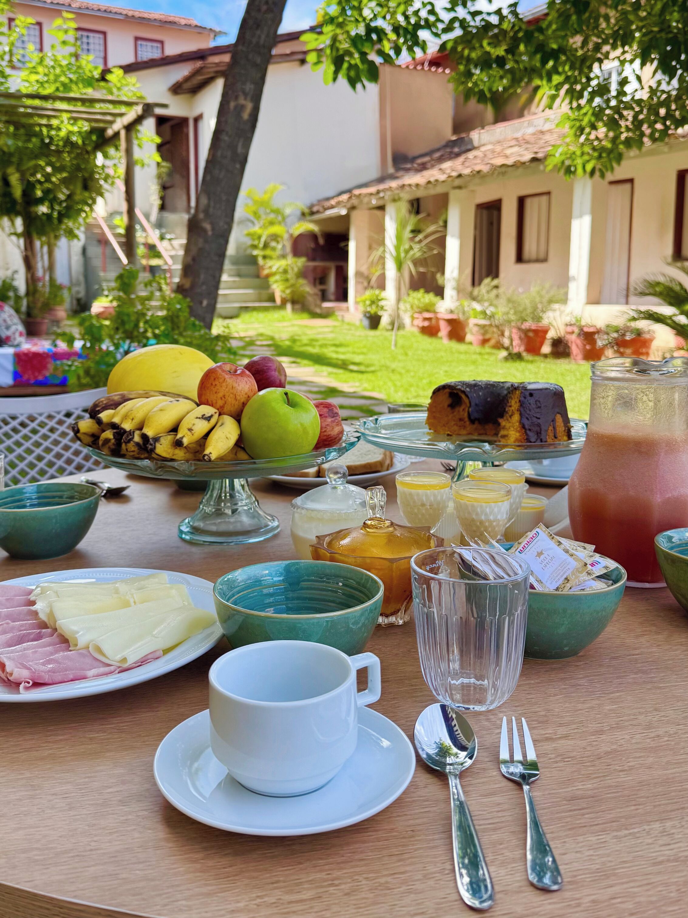 A breakfast table with fruits, pastries, and beverages, set outdoors with a view of a house and garden.