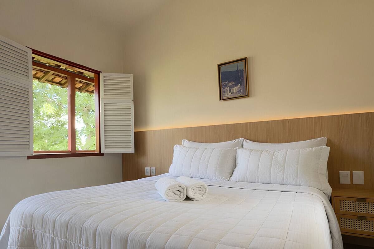 A neatly made bed with white linens and pillows, a wooden headboard, and a window with blinds.