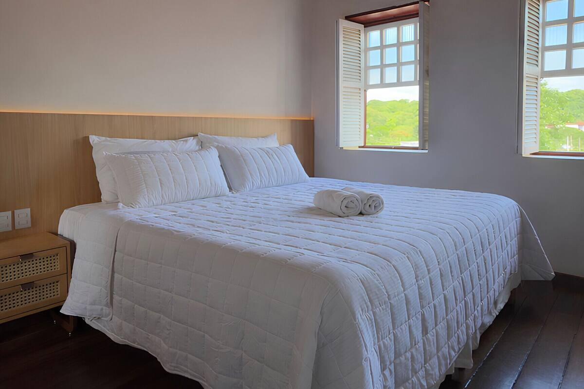 A neatly made bed with white bedding, a wooden headboard, and two windows with shutters.