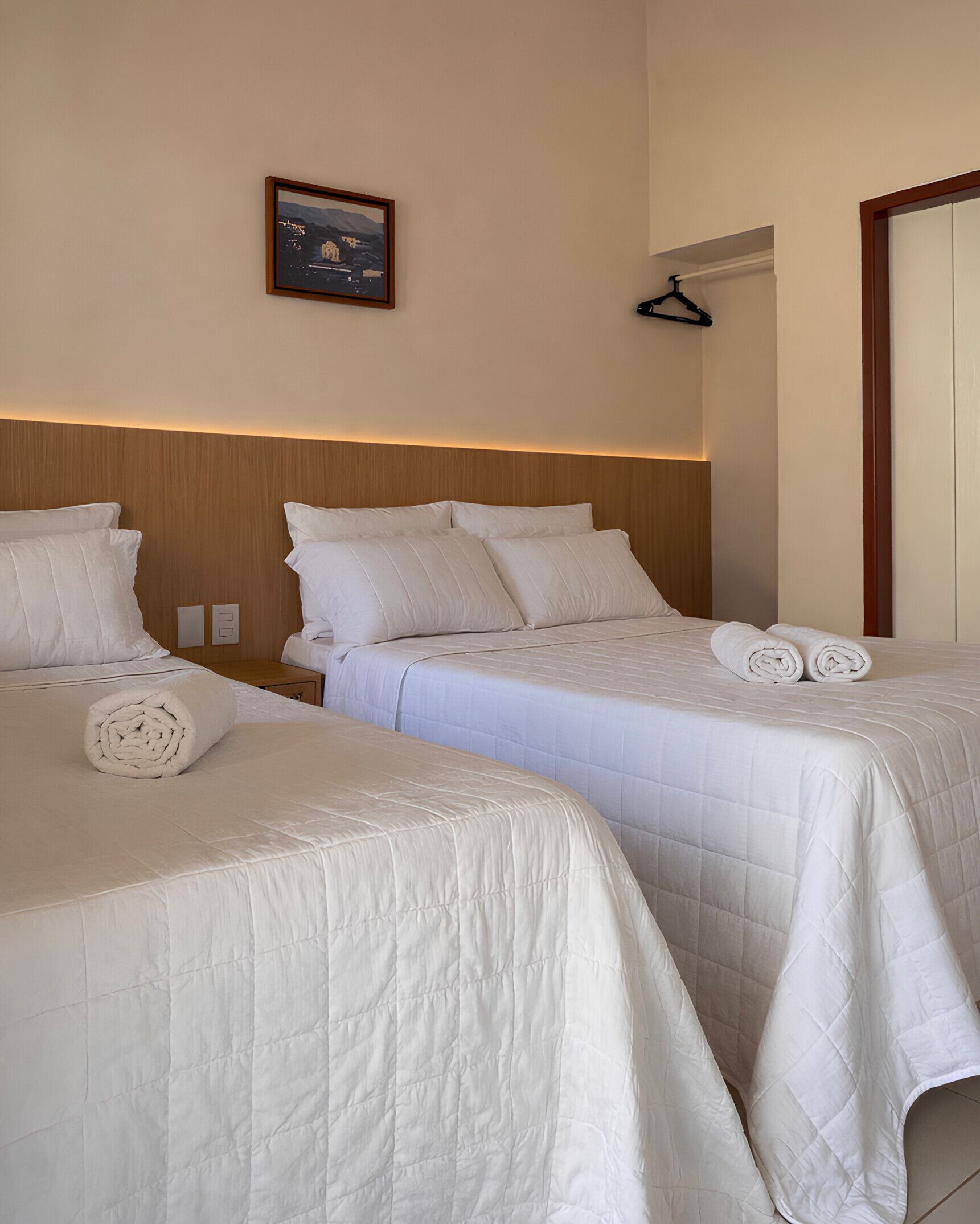 Ein Hotelzimmer mit zwei Betten, jeweils mit weißer Bettwäsche und Kissen. Über den Betten hängt ein gerahmtes Bild, eine Garderobe und eine Tür ist rechts zu sehen.