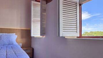 Quarto com cama, cabeceira de madeira, paredes brancas e janela com portaria.