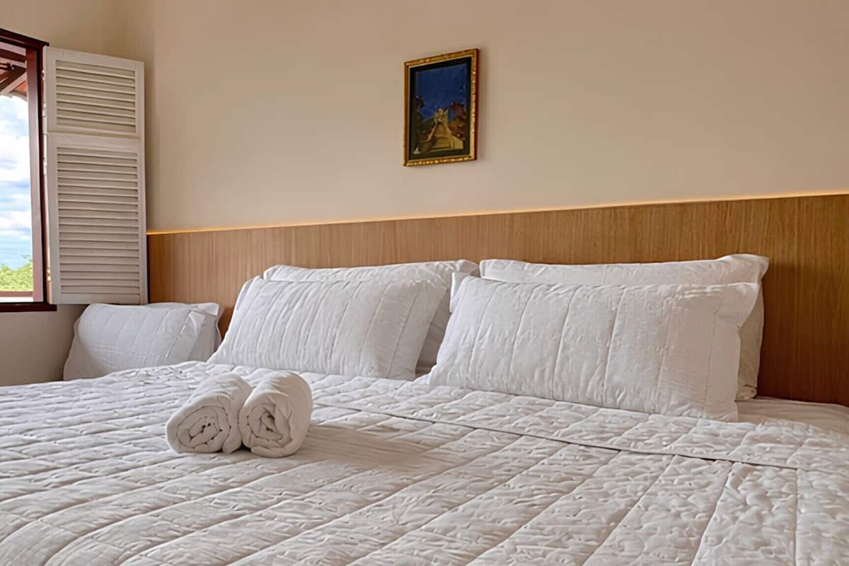 A neatly made bed with a quilted white comforter and rolled towels, a wooden headboard, and a framed picture on the wall.