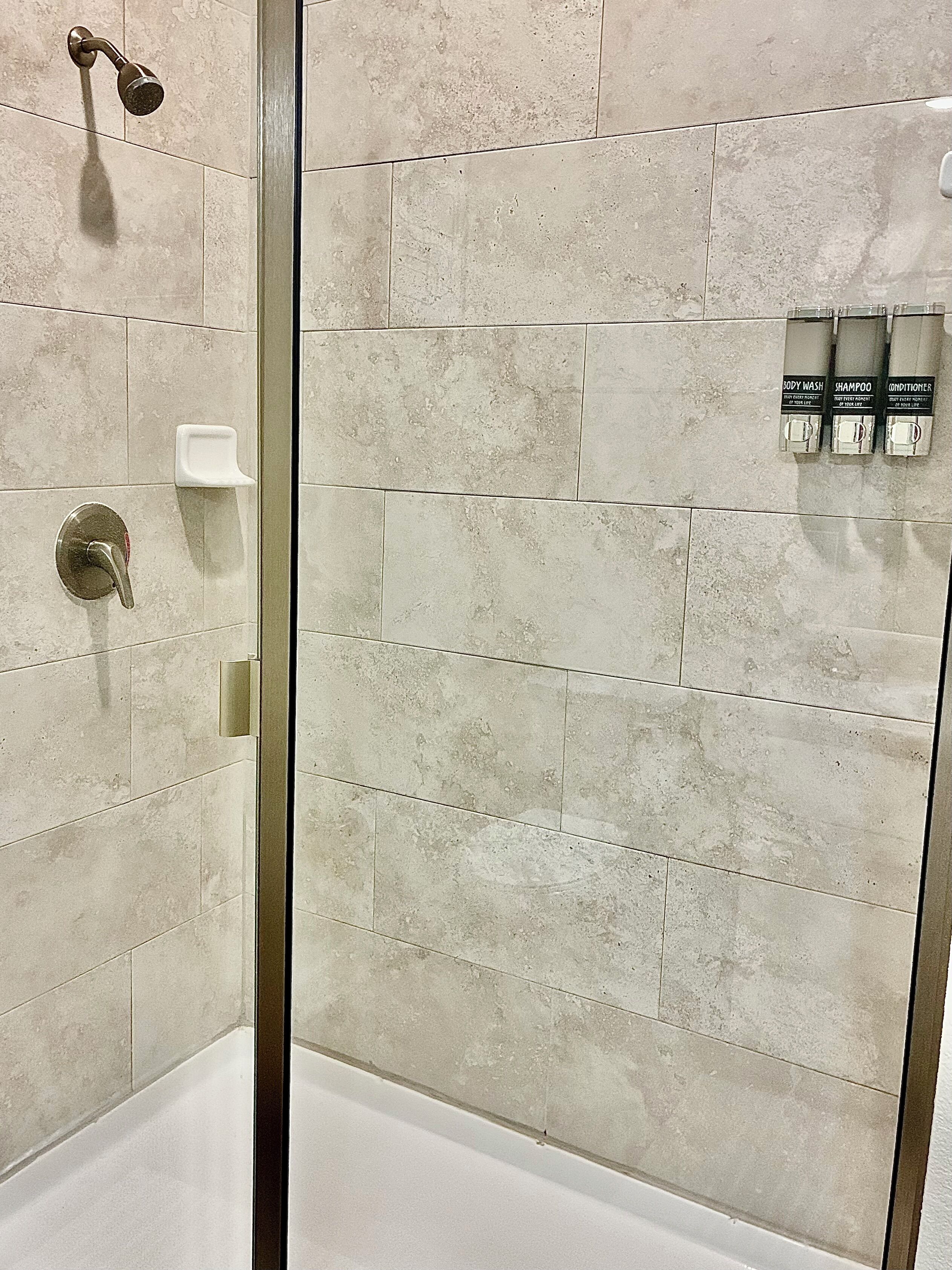 A shower area with a glass door, beige tiles, a handheld showerhead, and a soap dispenser.