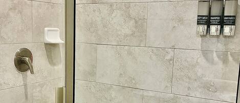 A shower area with a glass door, beige tiles, a handheld showerhead, and a soap dispenser.
