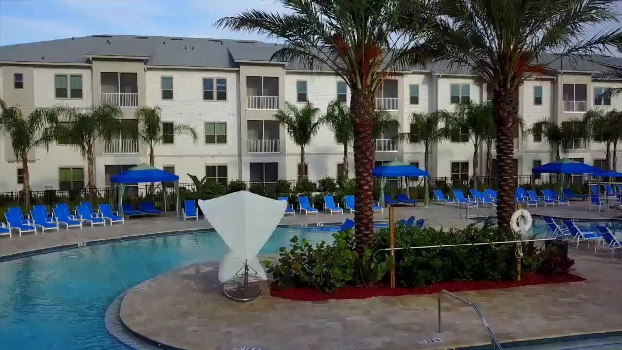 Un área de piscina con sillones reclinables azules y palmeras, rodeada por un edificio con balcones.