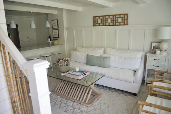 A modern living room with a white sofa, glass coffee table, and a kitchen area in the background.
