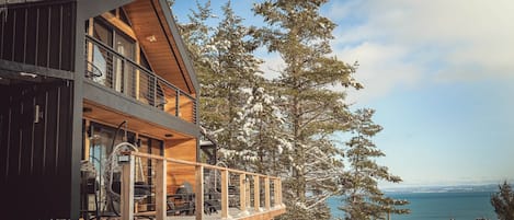 Una cabaña moderna con terraza de madera y amplios ventanales con vistas a un paisaje nevado y al agua.