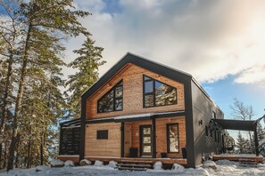 Une cabane moderne en bois, dotée d’une grande véranda à l’avant, de grandes fenêtres et d’un espace extérieur couvert.