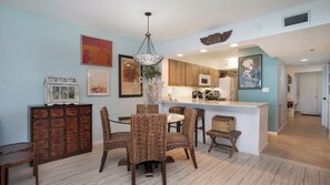 A dining area with a round table and wicker chairs, a kitchen with wooden cabinets, and a hanging light fixture. - 7th-Floor Beachfront Marco Island Condo with Panoramic Ocean & Sunset Views (Marco Island)