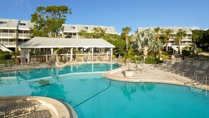 Ein Swimmingpool mit Liegestühlen und einer überdachten Sitzgruppe, umgeben von Palmen und einem Gebäude mit mehreren Balkonen.
