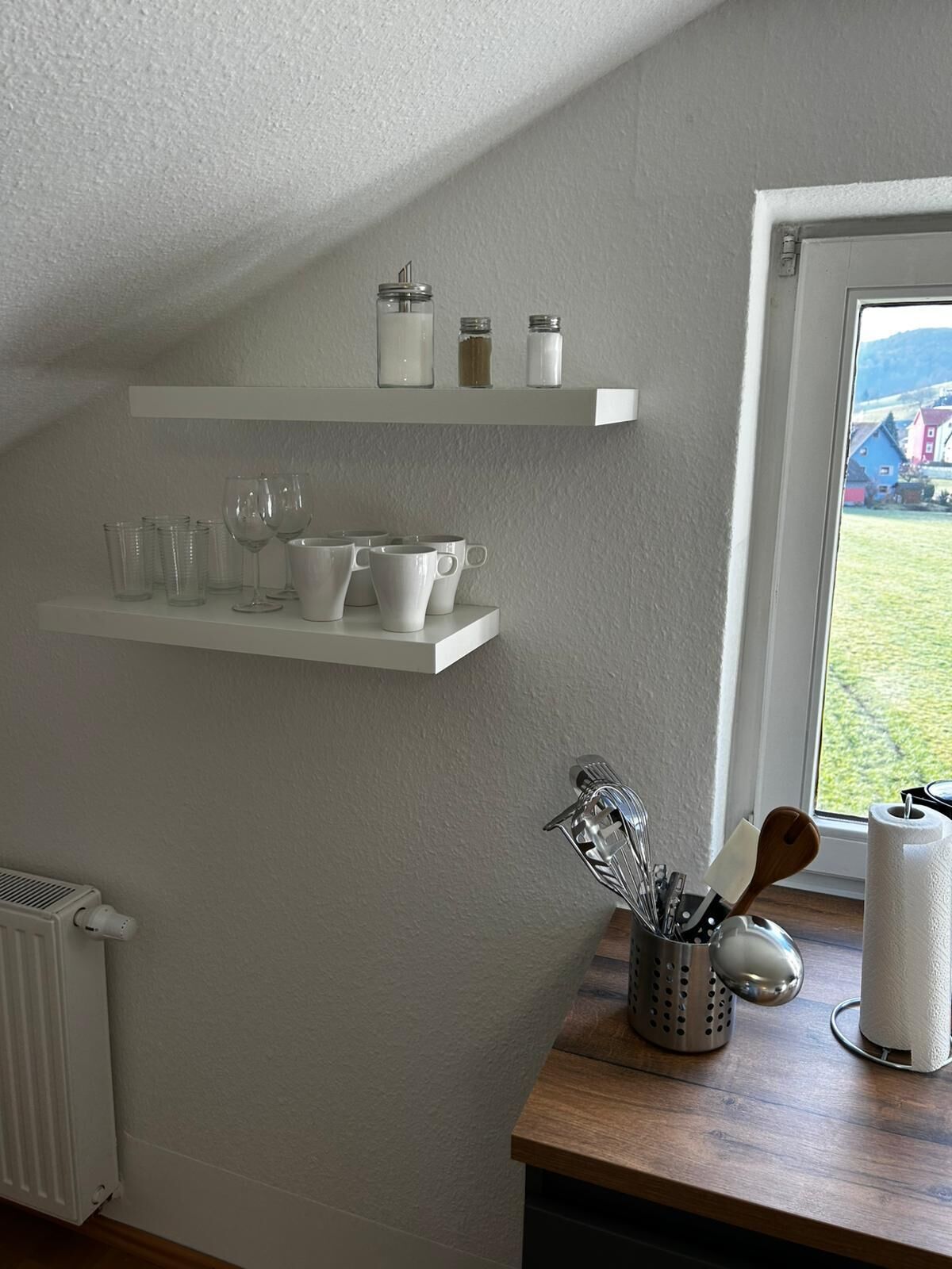 Une cuisine avec des étagères blanches où sont rangées des tasses et des verres, un salier, et une fenêtre donnant sur des maisons.