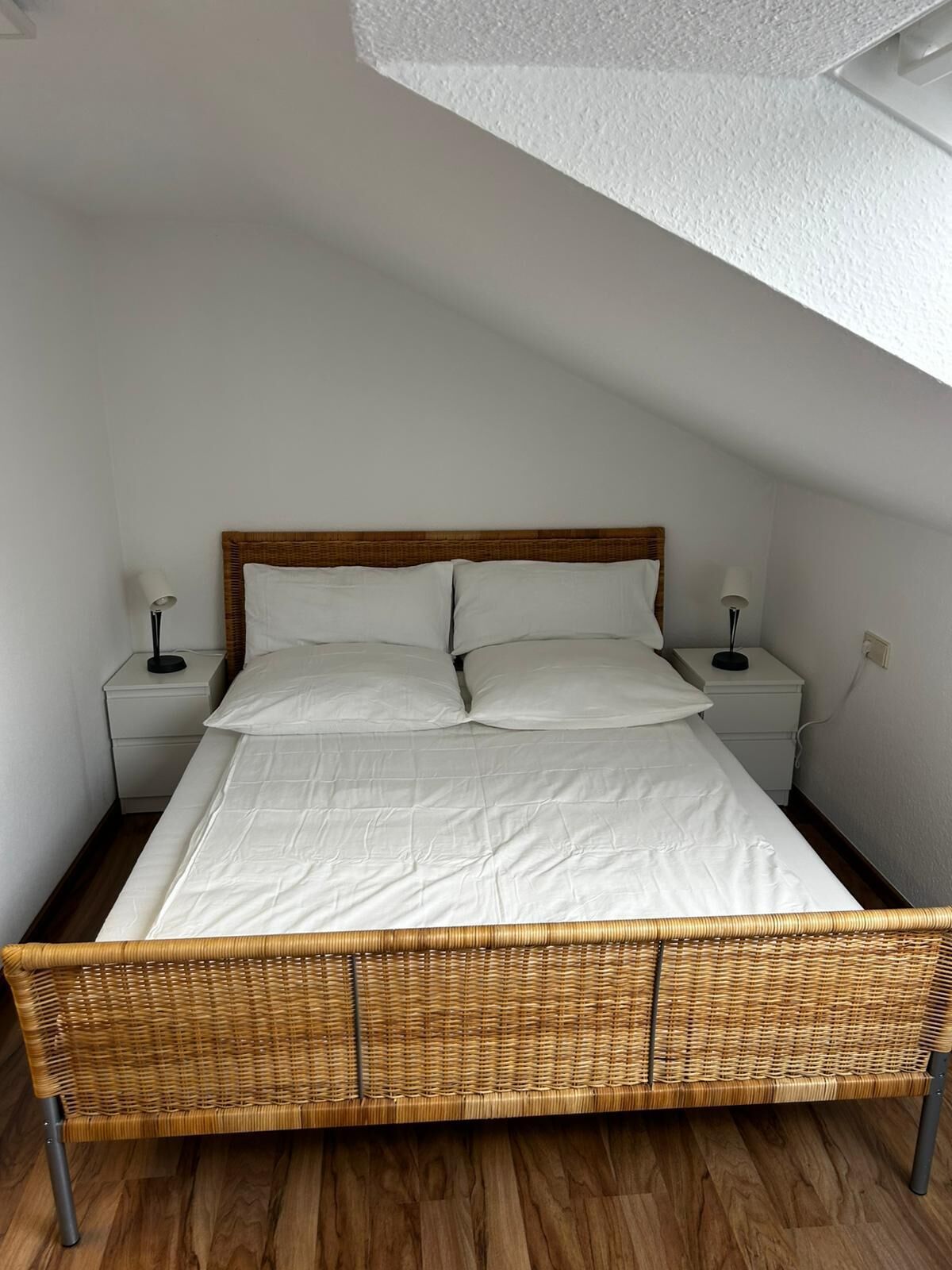 Uma cama de solteiro com cabeceira de madeira, roupa de cama branca, duas mesas de cabeceira com abajures e piso de madeira.