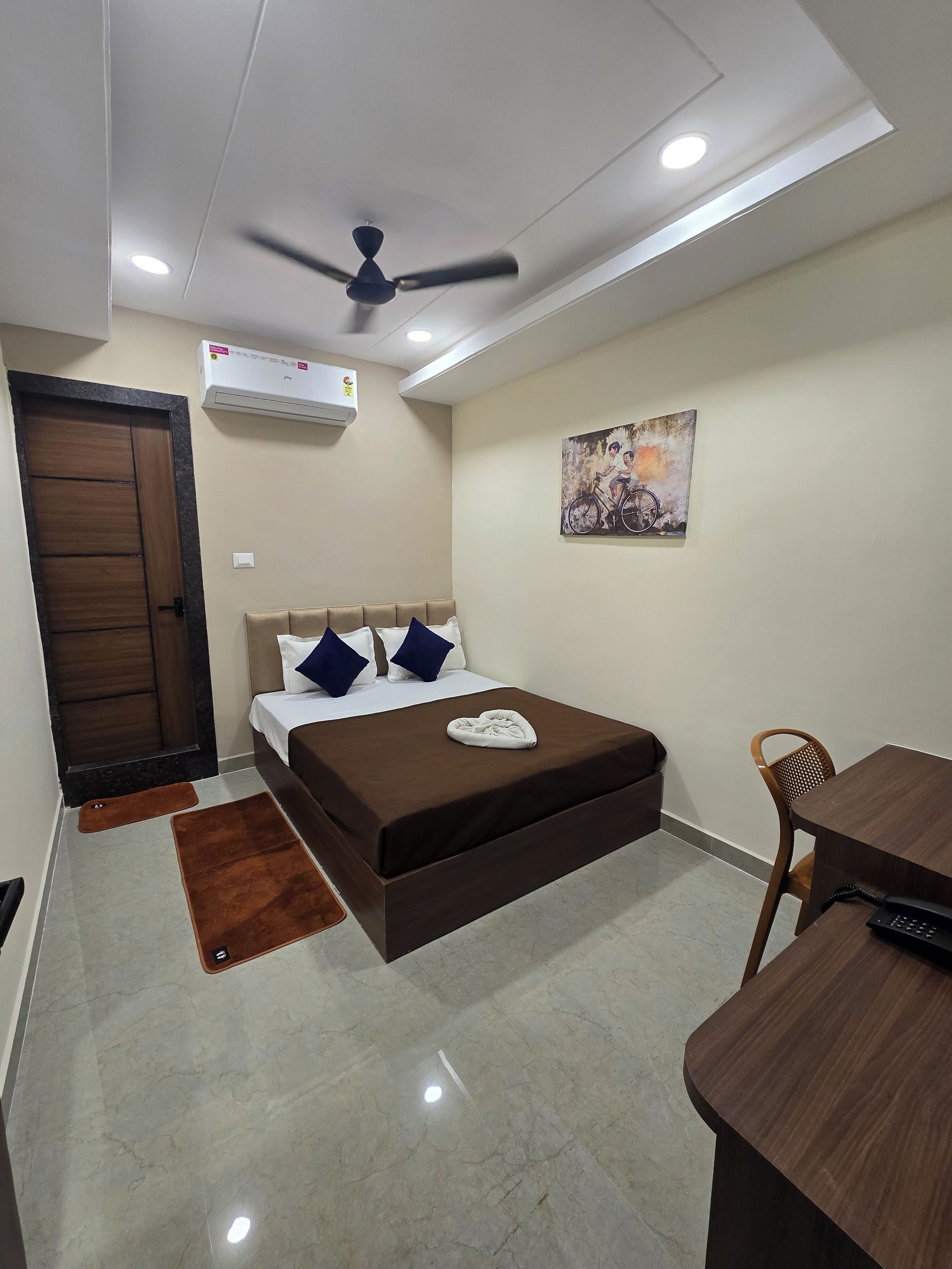 A hotel room with a bed, a desk, a chair, a ceiling fan, an air conditioning unit, and a painting on the wall.