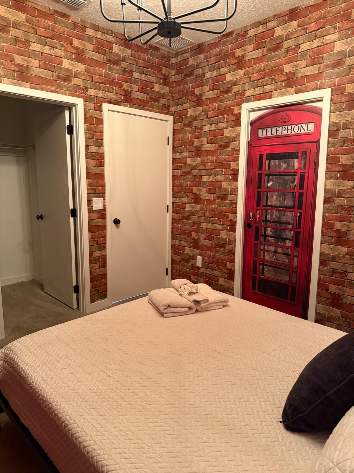 Un dormitorio con pared de ladrillo, cama con colcha color crema, puerta tipo cabina telefónica roja y ventilador de techo.