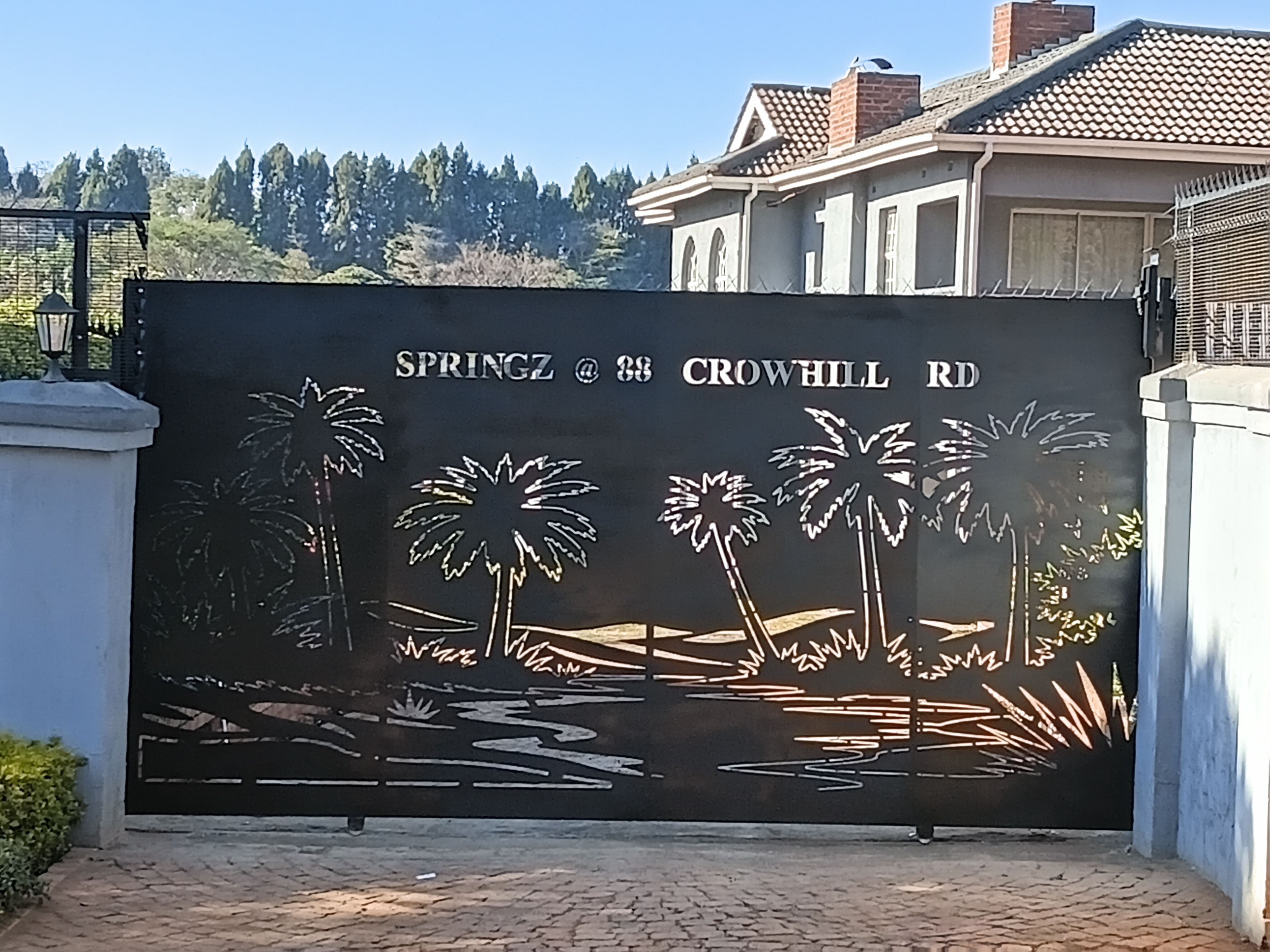 A metal gate with a landscape design, including palm trees and water, at the entrance to a property named SPRINGZ at 88 CROWHILL RD.