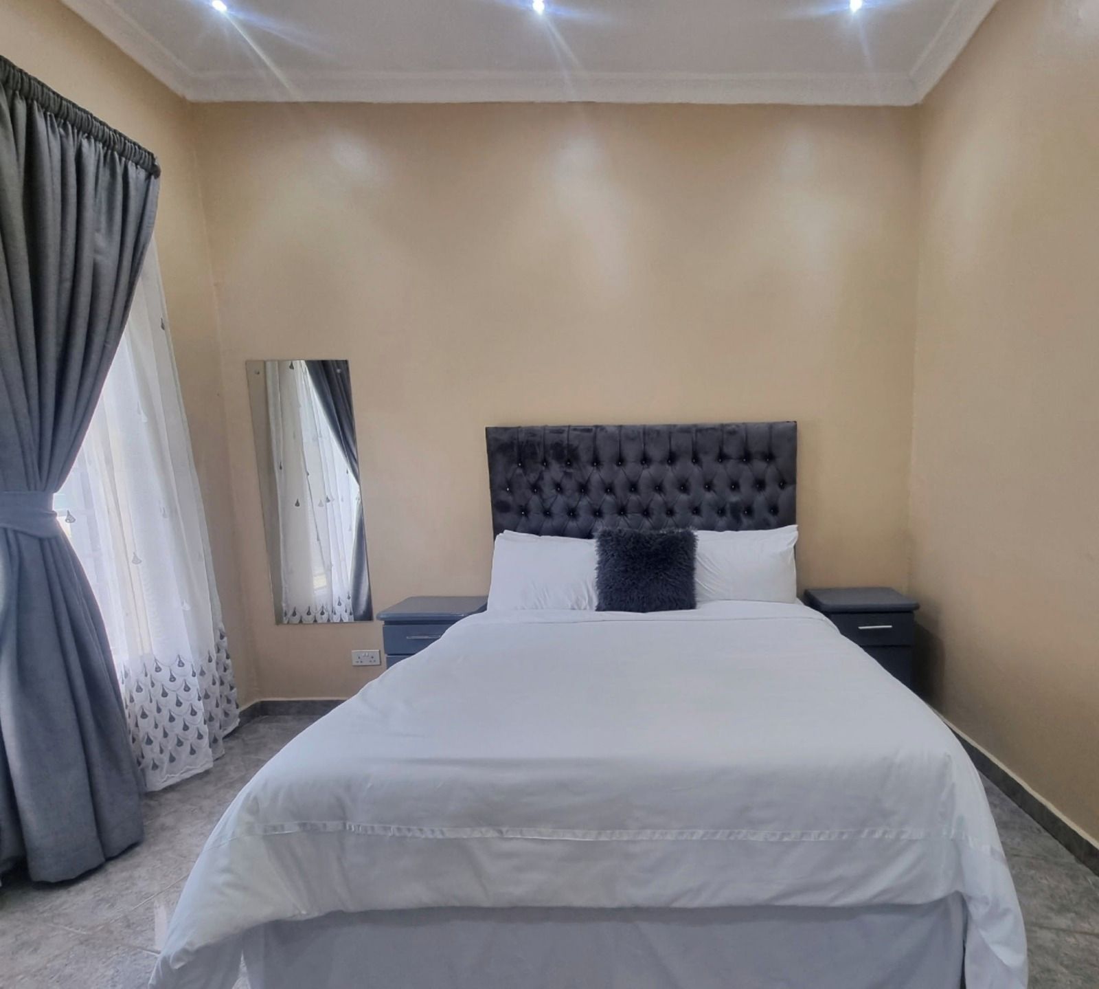 A hotel room with a large bed, a headboard, two bedside tables, a mirror, and a window with curtains.