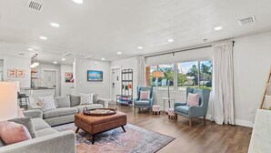 Un salón amplio con un sofá gris, un reposapiés marrón y una alfombra estampada. Hay dos sillones y una mesita redonda. La habitación tiene ventanas grandes con cortinas y vistas al exterior.