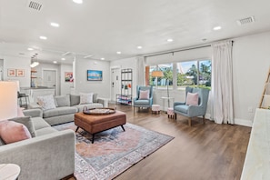 A spacious living room with a grey sofa set, a brown ottoman, and a patterned rug. There are two armchairs and a small round table. The room has large windows with curtains and a view of the outdoors. - Private Pool Oasis Near Anna Maria Island + Tiki Bar + Games + Sleeps 12 (Bradenton)