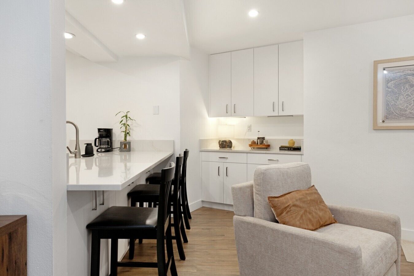 Une cuisine moderne avec des meubles blancs, un coin repas avec des chaises noires et un canapé gris agrémenté d’un coussin marron.