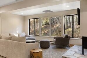 A modern living room with a beige sofa, a grey armchair, a round ottoman, and a wooden coffee table. Large windows provide a view of a forested area. - Fallridge Condo - Steps to Nordic Center & Bus (Vail)
