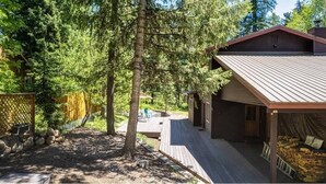 Une cabane dotée d’une terrasse en bois, entourée d’arbres, et d’un espace abrité où est entreposé du bois de chauffage.