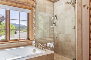 Une salle de bain dotée d’une grande fenêtre, d’une baignoire, d’une douche avec une paroi en verre et d’une étagère contenant des articles de toilette.