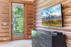 L’intérieur d’une cabane en rondins, avec une porte-fenêtre coulissante donnant sur une zone boisée, un téléviseur à écran plat fixé au mur et une commode en bois foncé.
