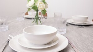 Een eettafel gedekt met witte borden, kommen en een vaas witte rozen.