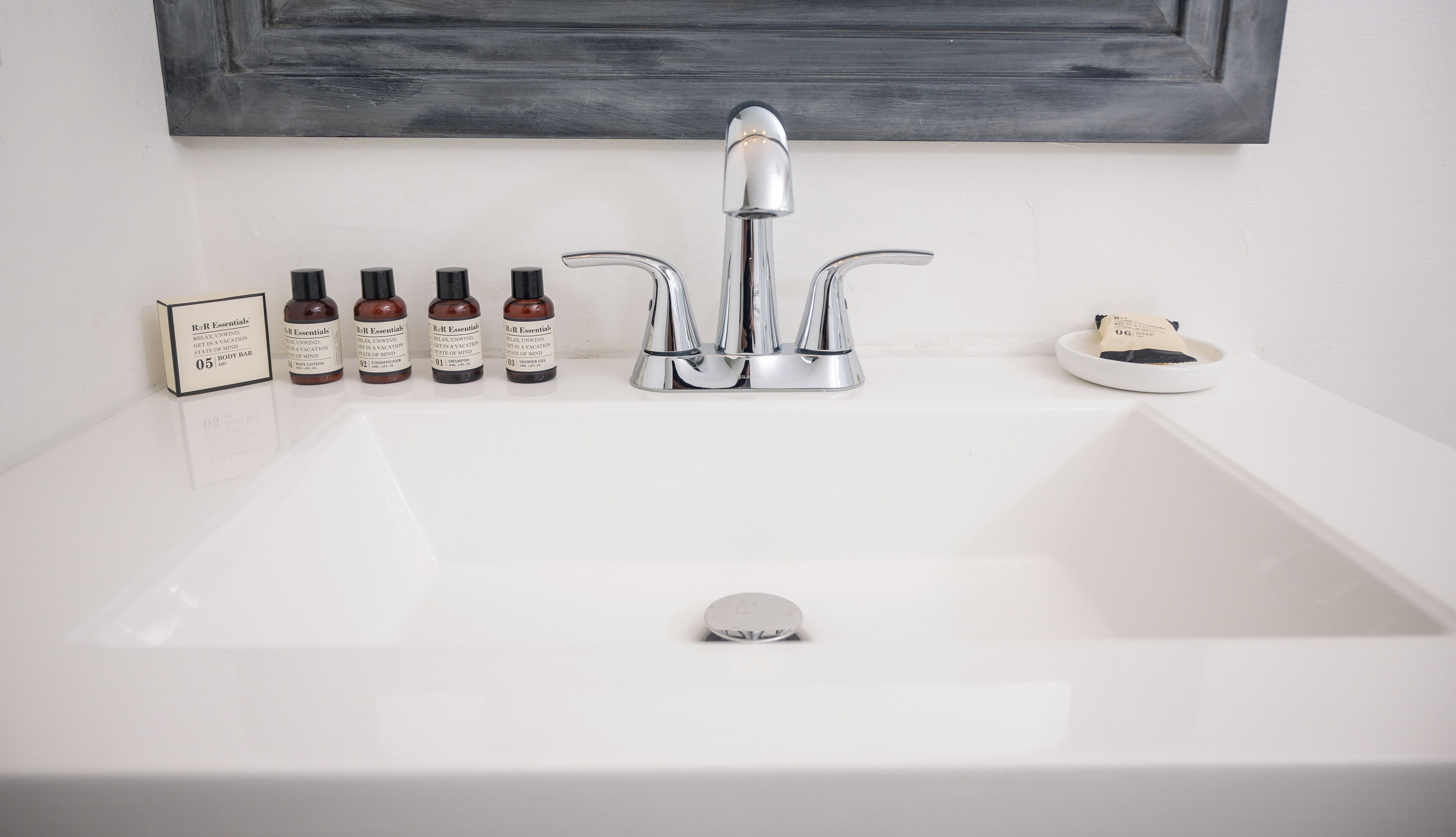 Un lavamanos blanco con una grifería moderna, cinco botellas de productos de cuidado corporal R&R Essentials y un jabonera con una pastilla de jabón.