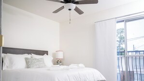 Een moderne hotelkamer met een groot bed, een plafondventilator, een nachtkastje met een lamp en uitzicht buiten door een groot raam.