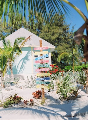Um poste de madeira com várias placas indicando direções para diferentes locais, como Praia, Bimini, Andros, Abaco, Eleuthera, Mayaguana, Exuma, Piscina e Inagua.