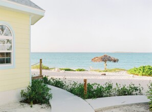 A beach view with a thatched umbrella, a sandy path, and a building with a window.