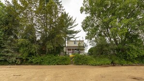 Une maison à deux étages avec un porche, entourée d’arbres denses et bordée d’un terrain sablonneux.