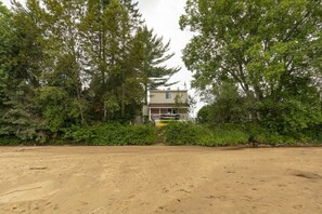 A two-story house with a porch, surrounded by dense trees and a sandy foreground.