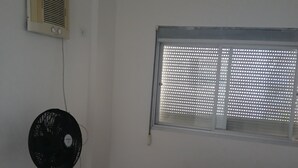 Ein Zimmer mit Bett, Standventilator, Fenster mit Jalousien und einer Wand-Klimaanlage.