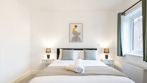 A neatly arranged bedroom with a bed, two bedside tables, a wall-mounted artwork, and a window with curtains. - Cozy 2-Bed near Heathrow/Thorpe Park/Legoland/ Long-Stay Friendly +Free Parking (Staines-upon-Thames)