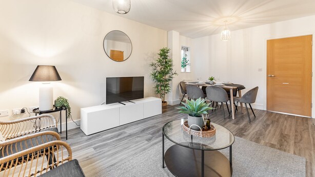 A modern living room with a flat-screen TV, a round mirror, a dining area with a table and chairs, and a wooden door. - Cozy 2-Bed near Heathrow/Thorpe Park/Legoland/ Long-Stay Friendly +Free Parking (Staines-upon-Thames)