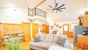 A spacious living area with a grey sofa, wooden walls, a ceiling fan, and a kitchen in the background.