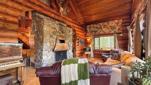 Interior de una cabaña de troncos con chimenea de piedra, un piano, un sofá y un televisor.