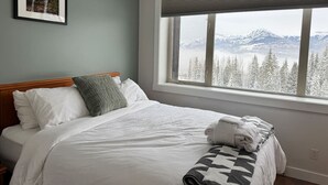A neatly made bed with white linens, a gray pillow, and a patterned blanket. A framed picture hangs on the wall above the bed. The room features a large window with a view of snow-covered mountains and trees.