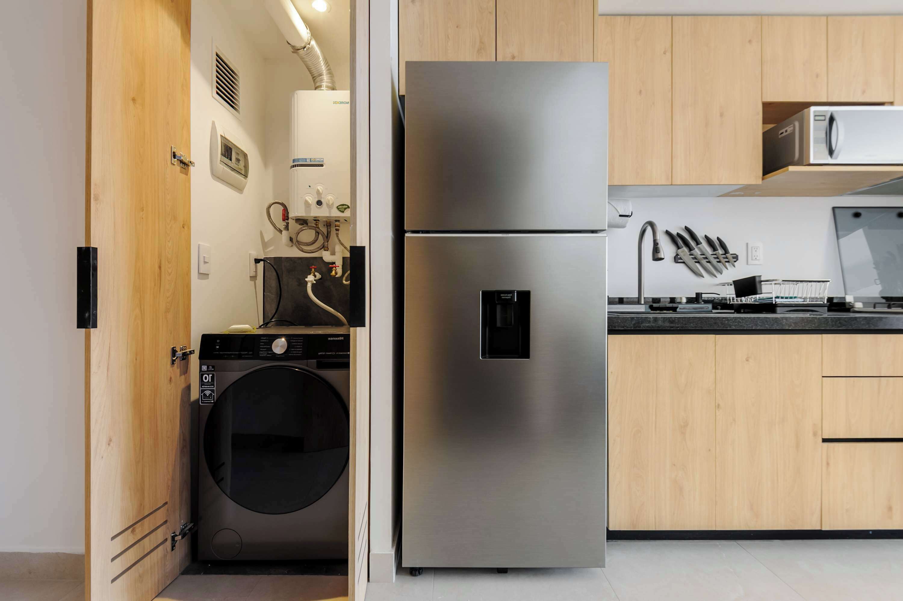 Een praktische keuken met een roestvrijstalen koelkast, een wasmachine, houten kastjes en een magnetron.