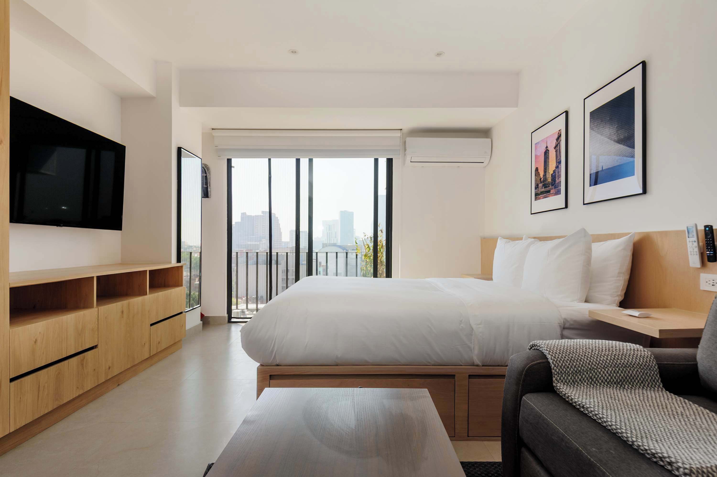 A modern hotel room with a large bed, a TV, a wooden cabinet, a sofa, and a balcony with a city view.