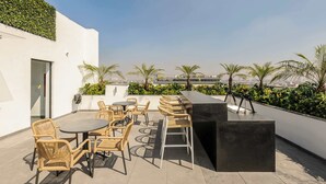 Een terras op het dak met rieten meubels, een bar en uitzicht op palmbomen en gebouwen.