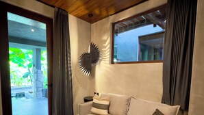 Ein modernes Wohnzimmer mit einem weißen Sofa, einem schwarzen Hocker und einer Wanddekoration. Ein großes Fenster mit Vorhängen lässt natürliches Licht herein und bietet Blick ins Grüne.